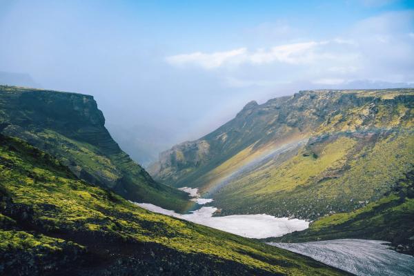 A vibrant green canyon with snow on the valley floor and a rainbow arching across.