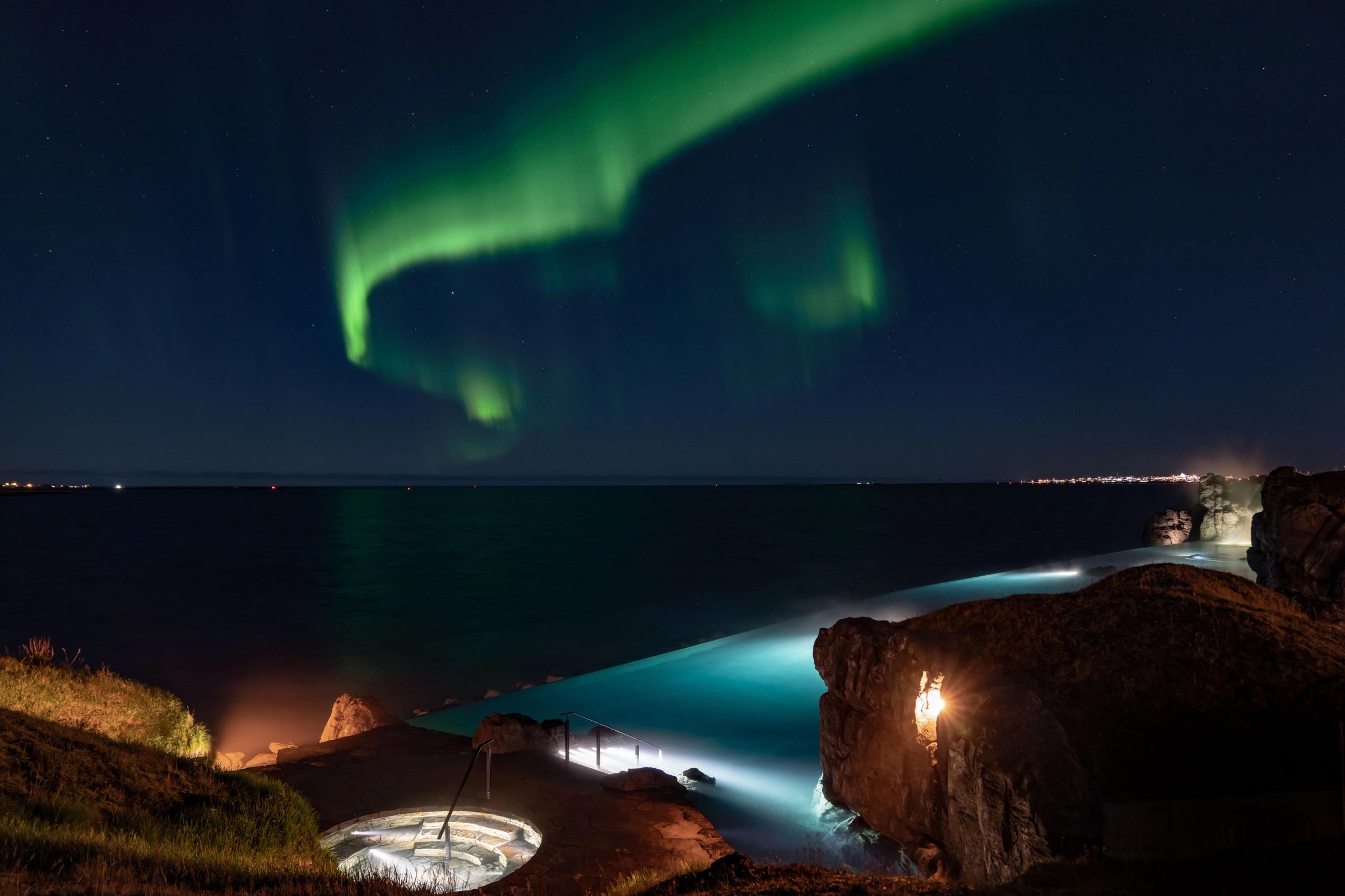 Green Northern Lights glow above a coastal infinity pool, ocean, and a circular sunken lounge at night.