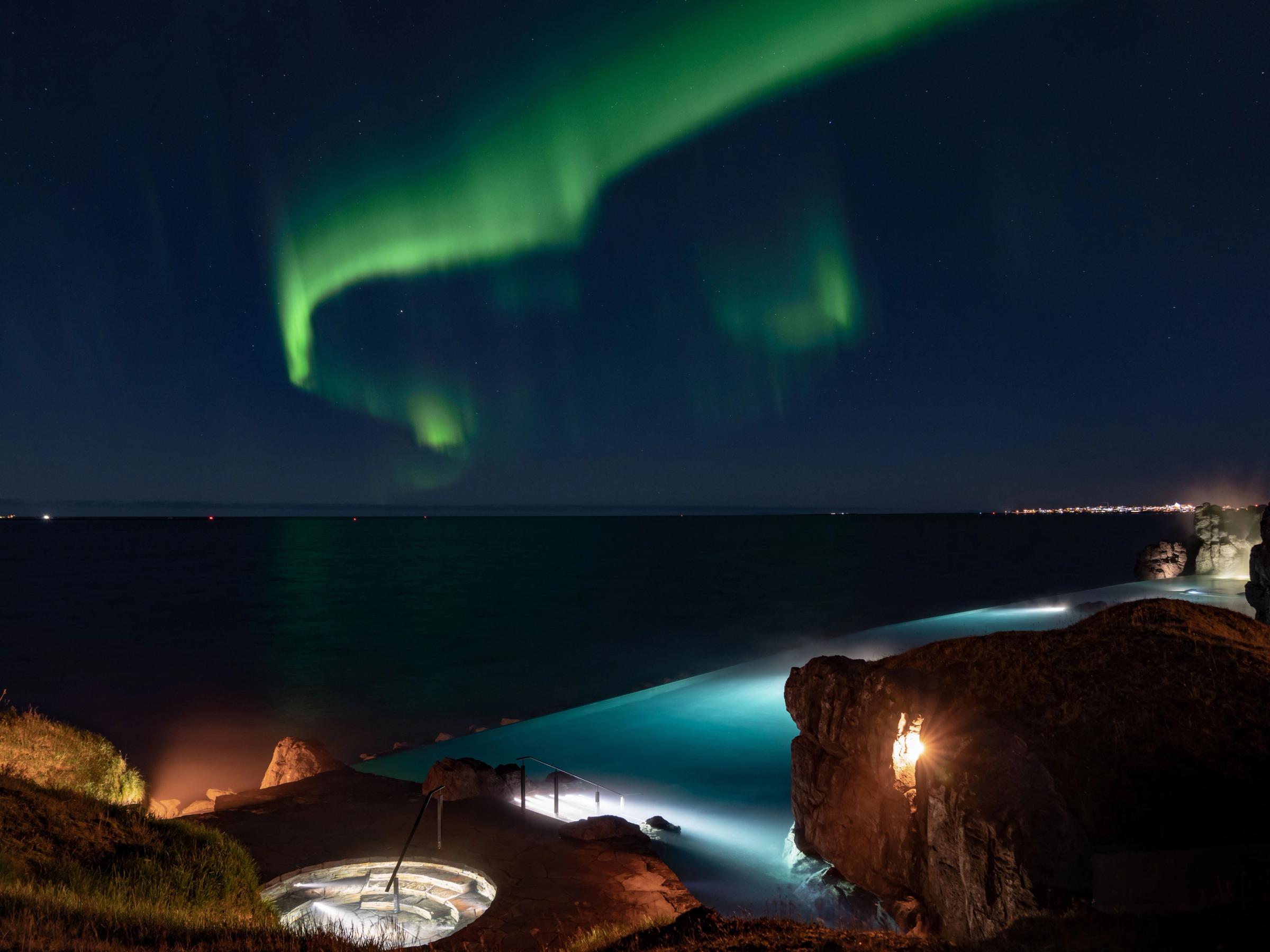 Green Northern Lights glow above a coastal infinity pool, ocean, and a circular sunken lounge at night.