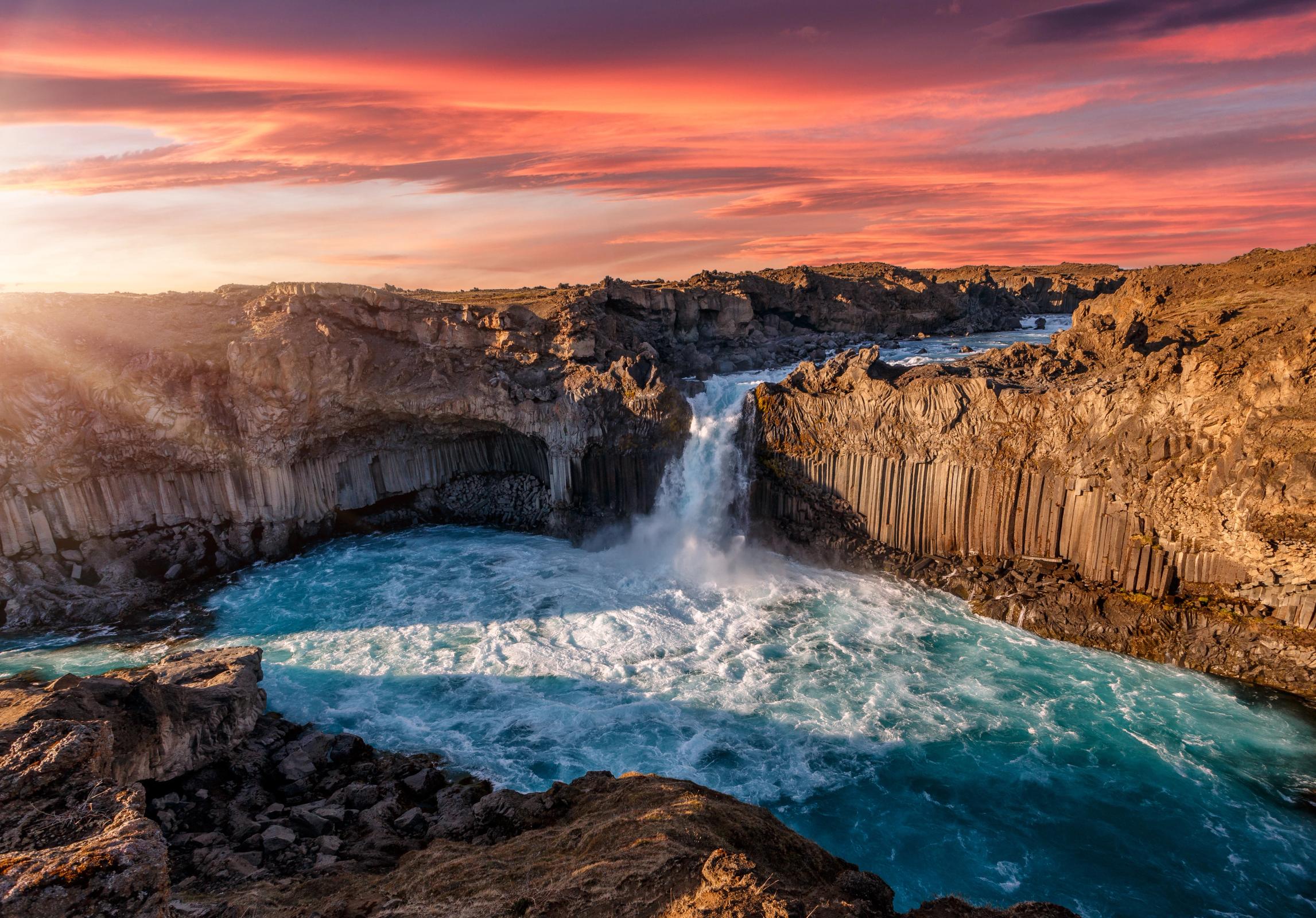Una poderosa cascada se precipita en un vibrante río turquesa rodeado de acantilados de columnas de basalto bajo un dramático cielo anaranjado y rojo al atardecer.