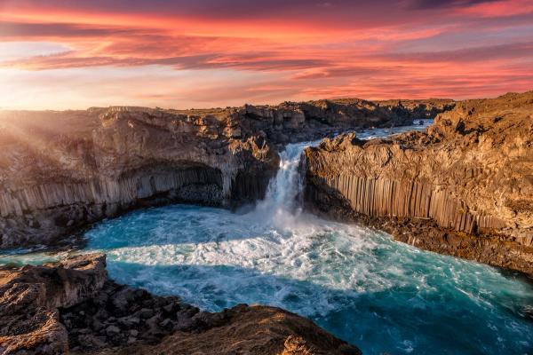 A powerful waterfall plunges into a vibrant turquoise river surrounded by basalt column cliffs under a dramatic orange and red sunset sky.