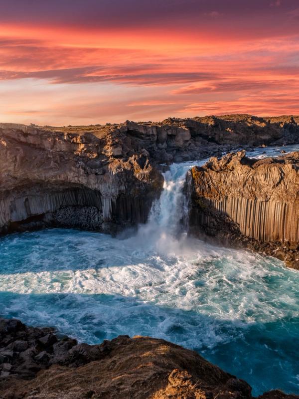 A powerful waterfall plunges into a turquoise river, flanked by columnar basalt cliffs under a vibrant sunset sky.