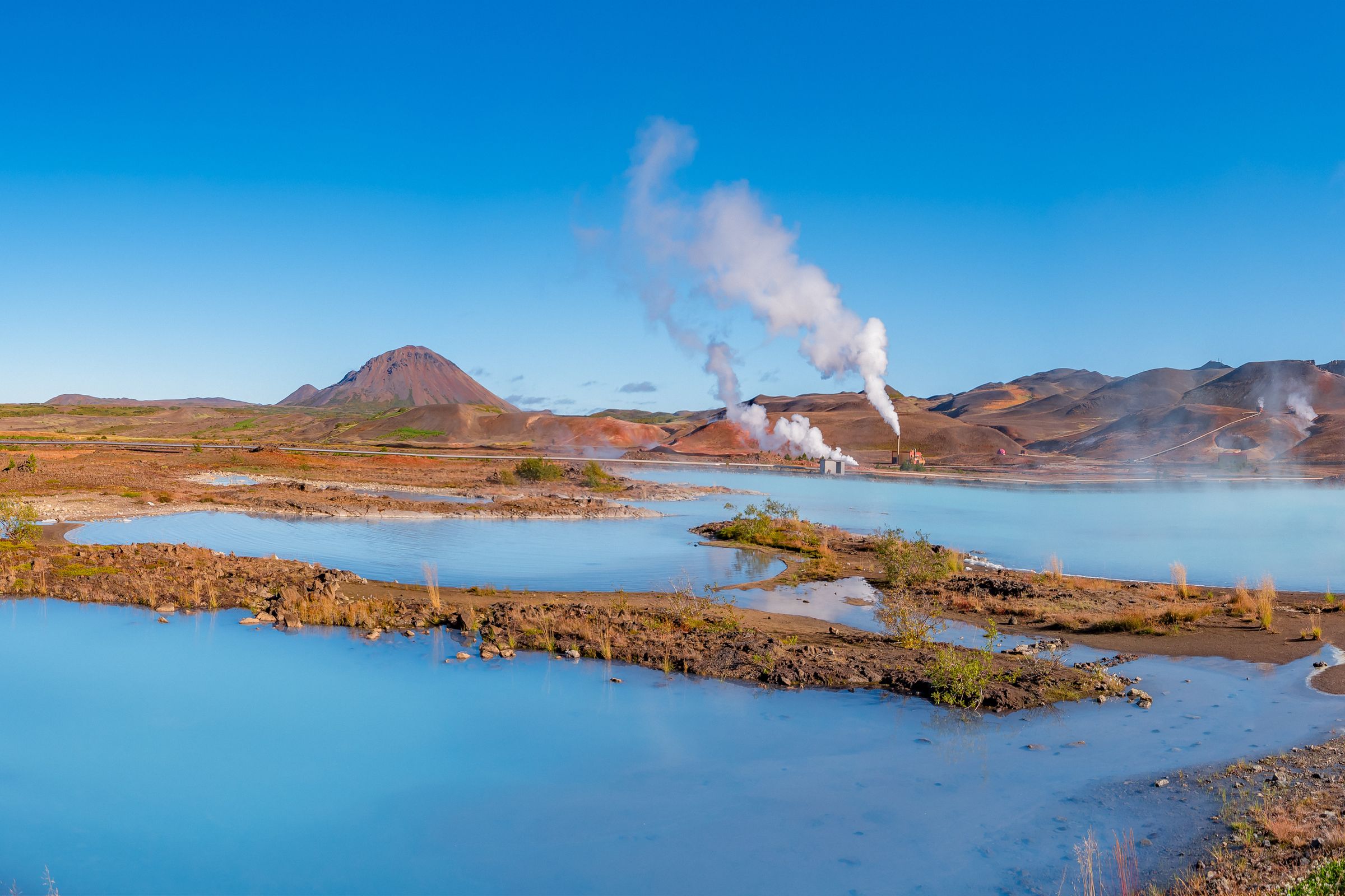 Vista panorámica de un lago geotérmico de un azul intenso, con vapor elevándose del agua y colinas de tonos rojizos y marrones alrededor, bajo un cielo despejado.