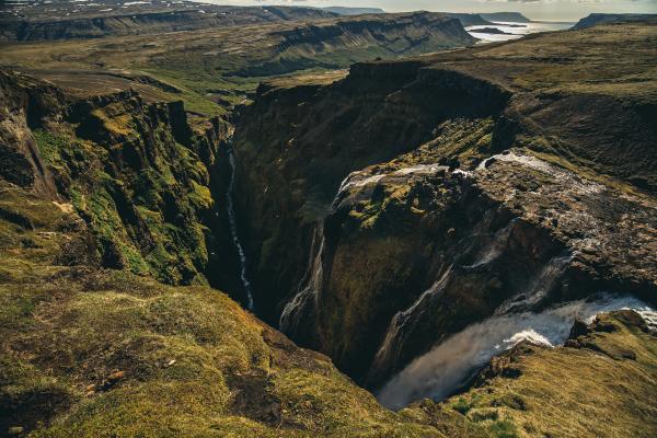 A powerful waterfall plunges into a deep, mossy canyon with a river flowing through, set in a vast, rugged landscape.