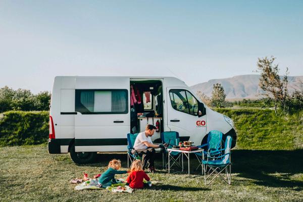 A white Go Campers campervan parked in a grassy field with a man and two children relaxing outside.