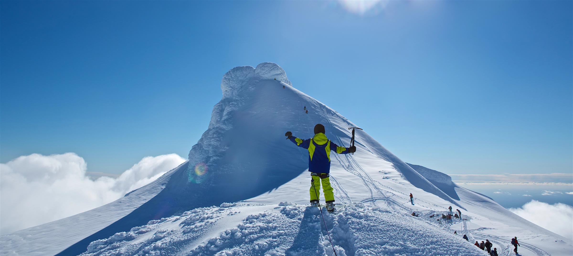 A person in yellow and blue celebrates on a snowy mountain peak, holding an ice axe with arms outstretched under a clear blue sky.