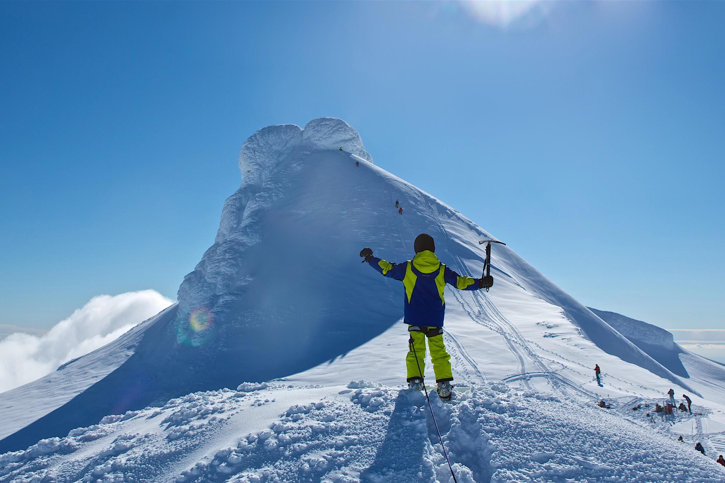 A person in yellow and blue celebrates on a snowy mountain peak, holding an ice axe with arms outstretched under a clear blue sky.