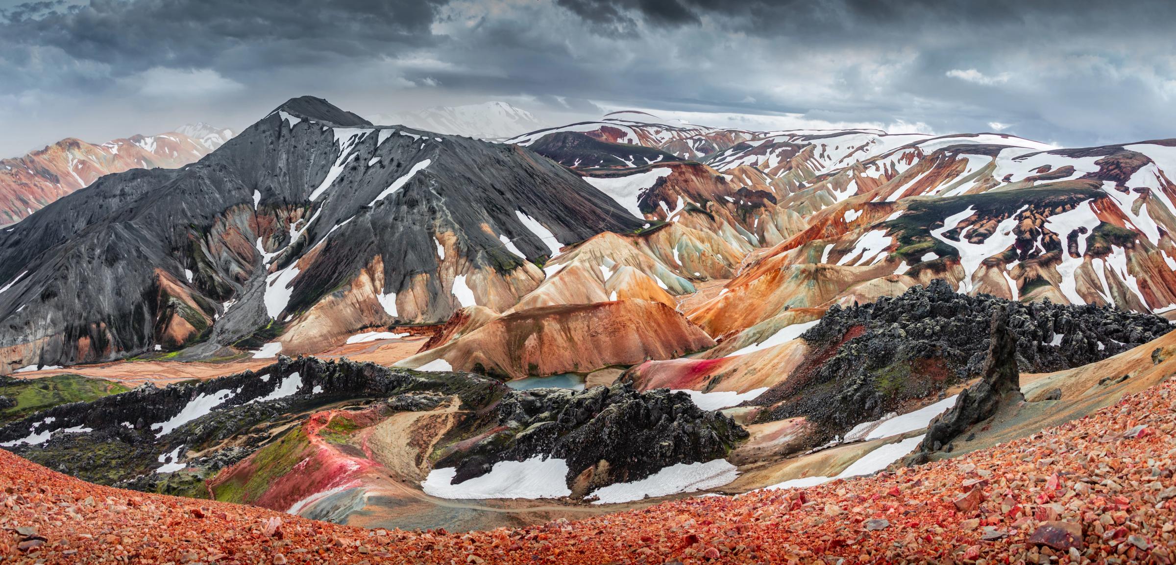 Vista panorámica de coloridas montañas volcánicas con parches de nieve, un pequeño lago y rocas de lava oscura bajo un cielo nublado.