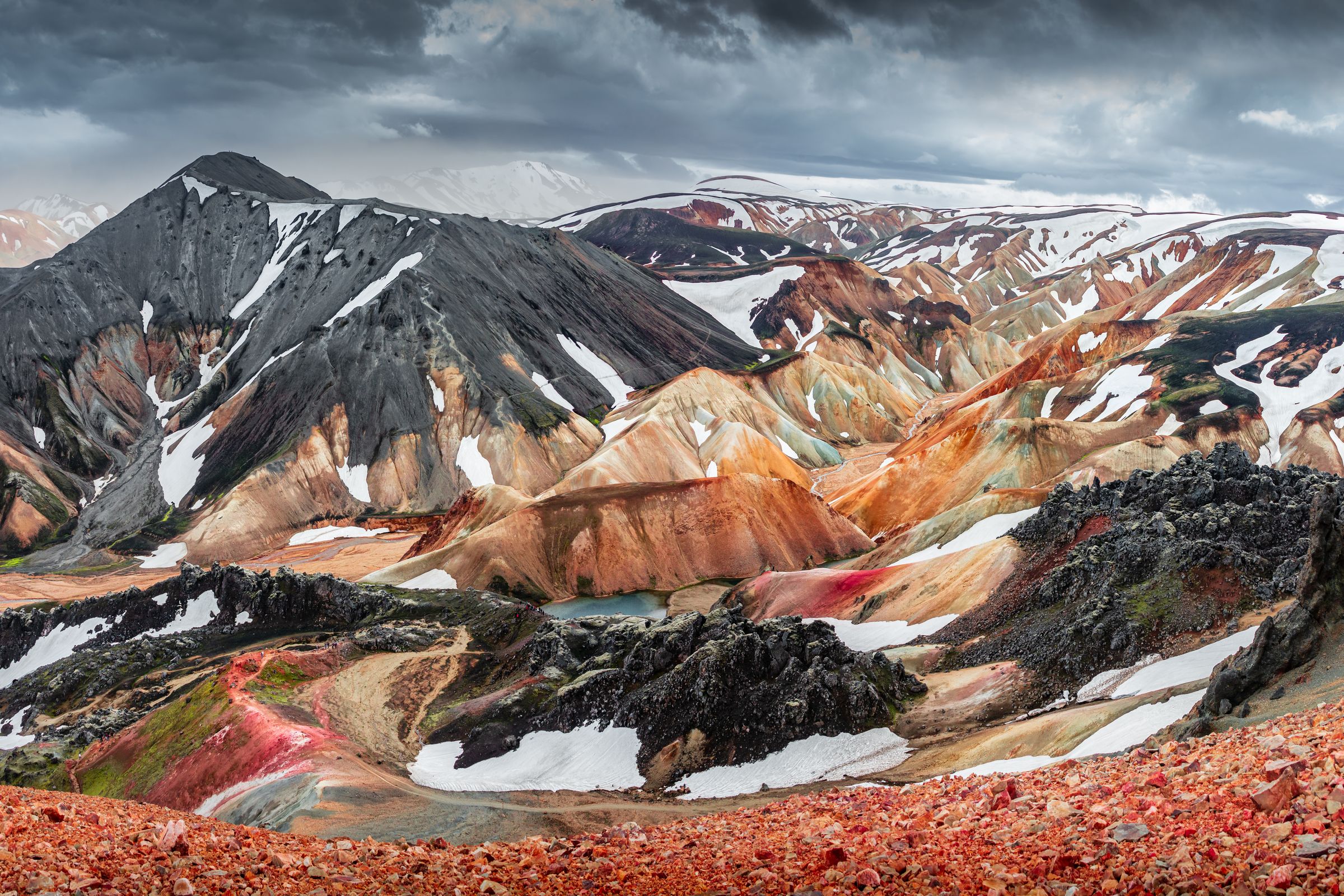 Vista panorámica de coloridas montañas volcánicas con parches de nieve, un pequeño lago y rocas de lava oscura bajo un cielo nublado.