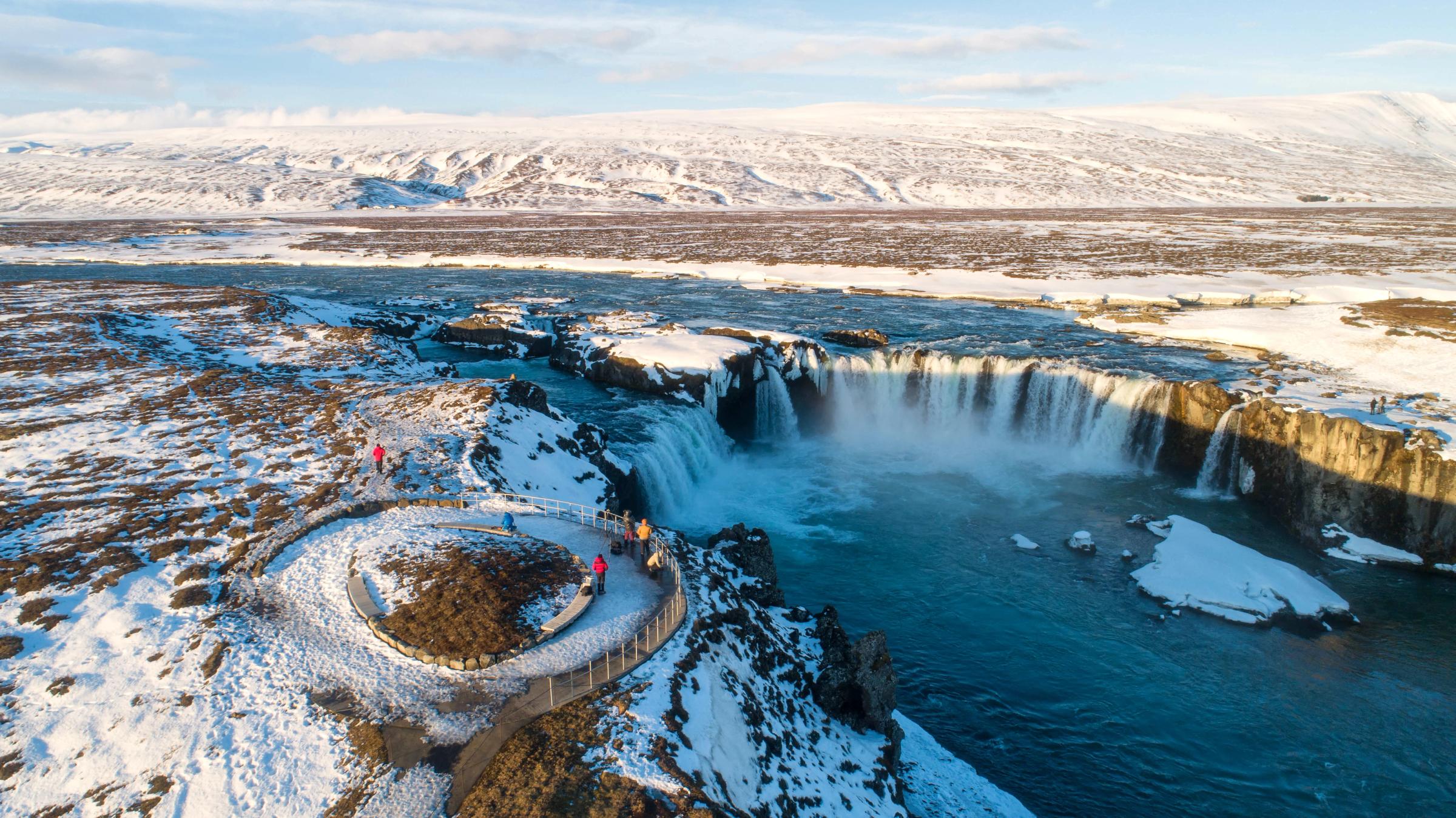 Aerial view of a wide waterfall in a snowy landscape, with people on a viewing platform.