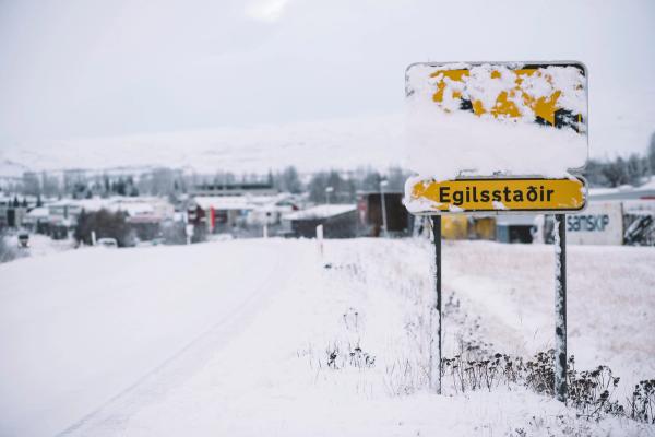 A snow-covered road sign for "Egilsstaðir" stands beside a snowy road leading to a town.