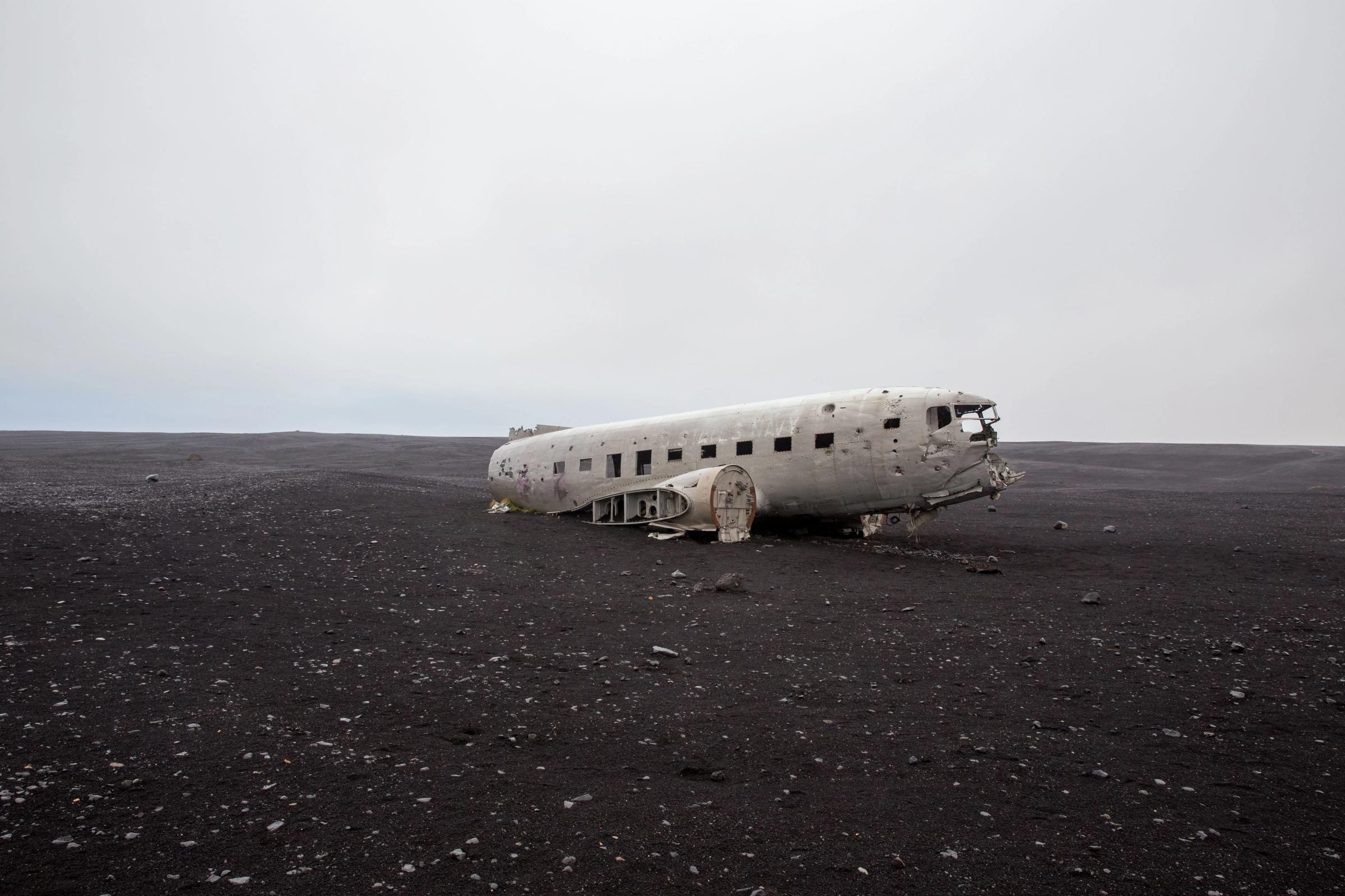 A white plane wreck sits on a vast black sand plain under a grey sky.