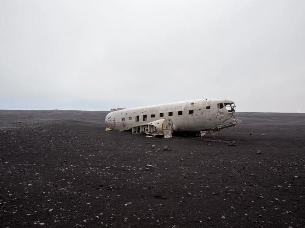 Wrecked airplane fuselage on a vast black sand plain under an overcast sky.