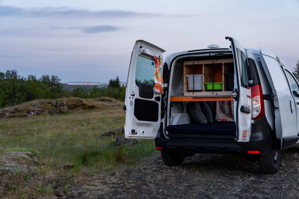 A white camper van with its rear doors open, revealing a bed and storage inside, parked on a dirt path in a rocky, grassy landscape at dusk.
