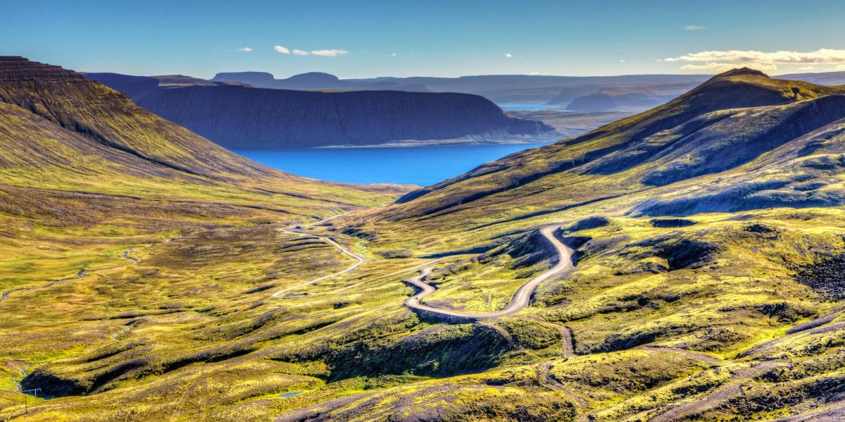 an aerial view of a mountain valley with a road going through it and a lake in the background in westfjords iceland.