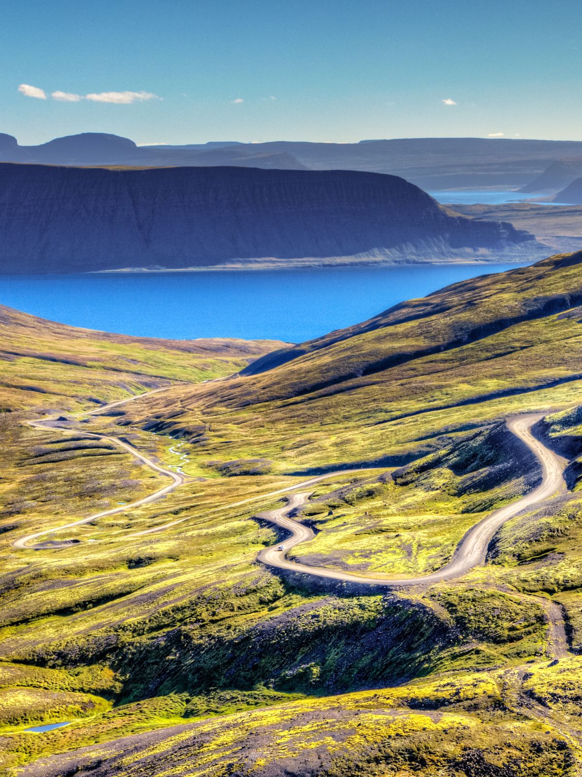 an aerial view of a mountain valley with a road going through it and a lake in the background in westfjords iceland.