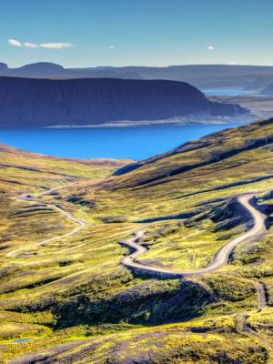an aerial view of a mountain valley with a road going through it and a lake in the background in westfjords iceland.