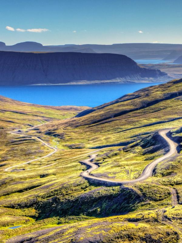 an aerial view of a mountain valley with a road going through it and a lake in the background in westfjords iceland.