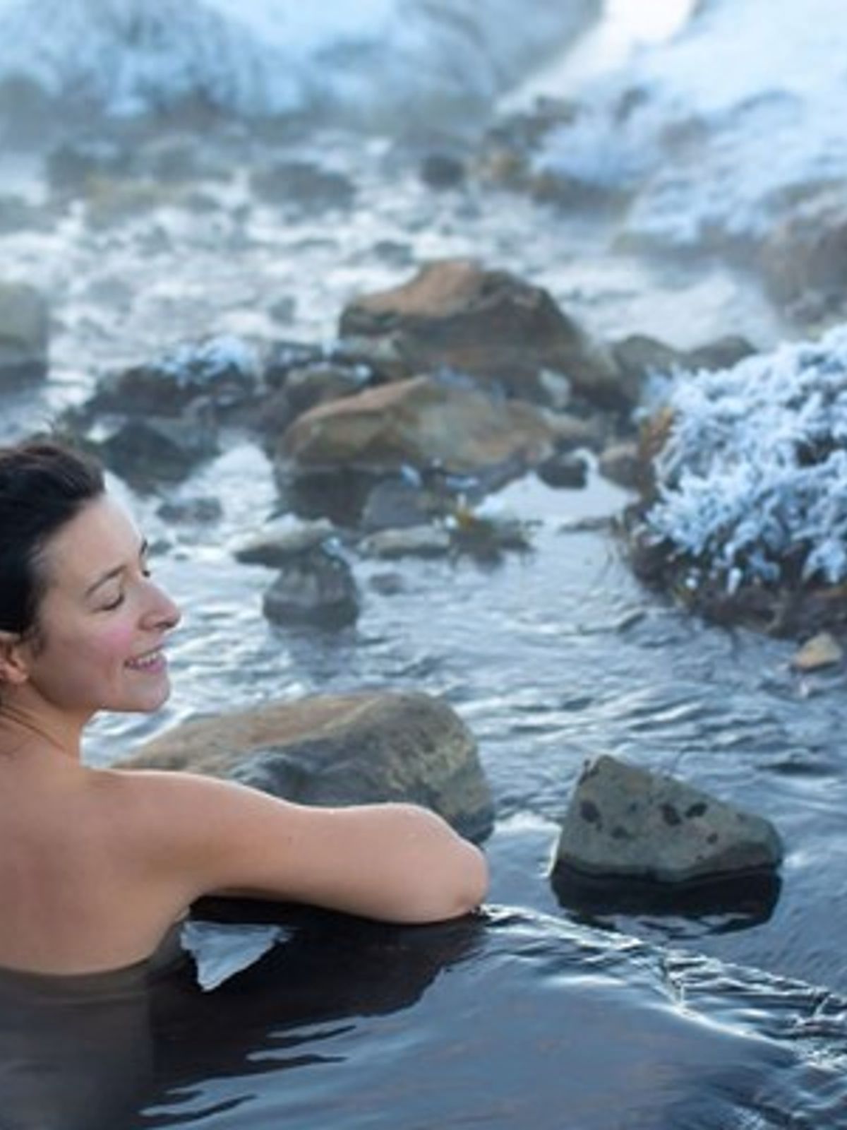 Smiling woman soaking in a warm outdoor stream with snowy banks.