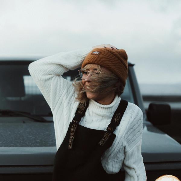 A smiling woman in a beanie and overalls, with windblown hair, leans against a dark vehicle.
