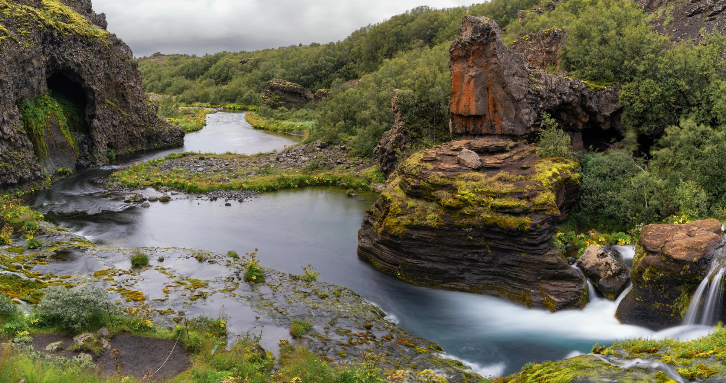 A winding river flows through a green, rocky valley featuring a cave, lush trees, and small waterfalls.