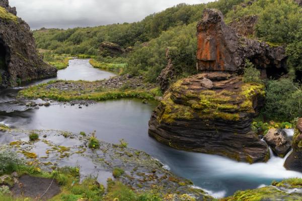A winding river flows through a green, rocky valley featuring a cave, lush trees, and small waterfalls.