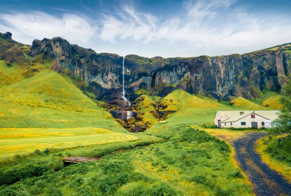 A waterfall cascades down dark cliffs in a lush green valley, with a white building and dirt road to the right.