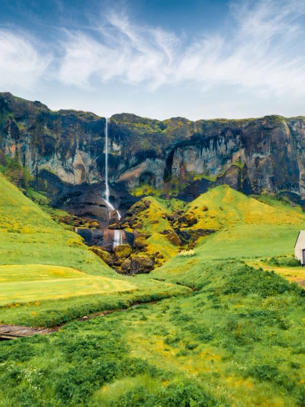 A waterfall cascades down dark cliffs in a lush green valley, with a white building and dirt road to the right.
