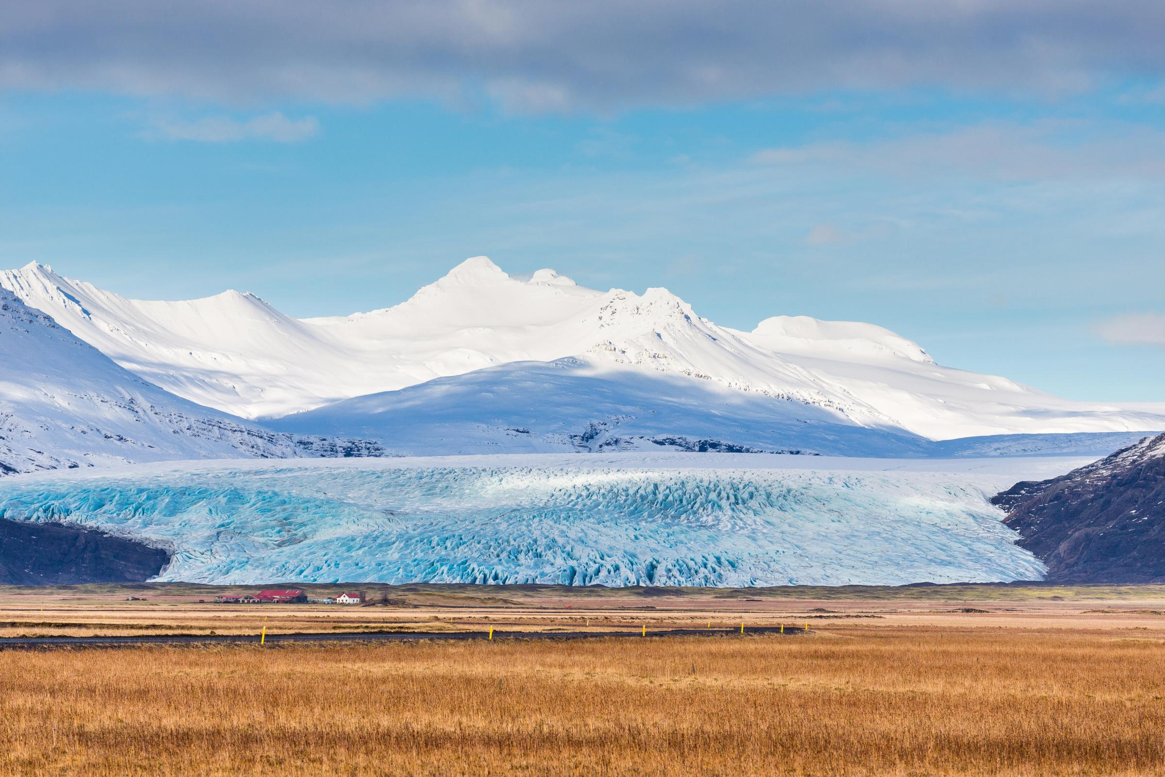 a glacier tongue with a snow covered mountain in the background and a brown grass field in the foreground 
