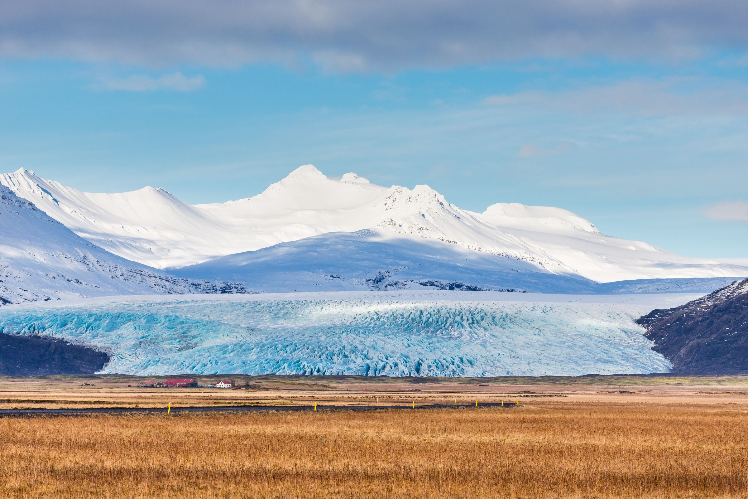 a glacier tongue with a snow covered mountain in the background and a brown grass field in the foreground