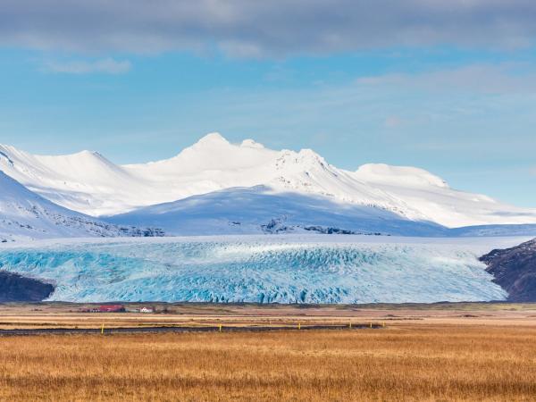 A vibrant blue glacier stretches across the landscape with snowy mountains behind it and a brown grassy field in the foreground.
