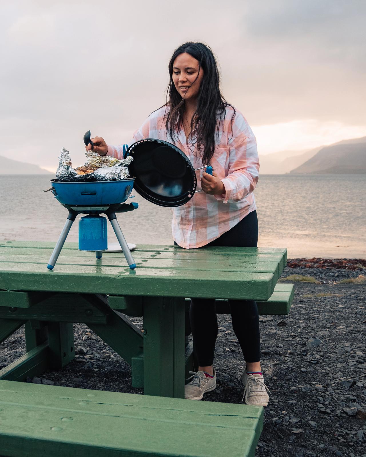 A woman lifts the lid of a portable blue grill cooking foil-wrapped food, on a green picnic table by a lake with mountains.
