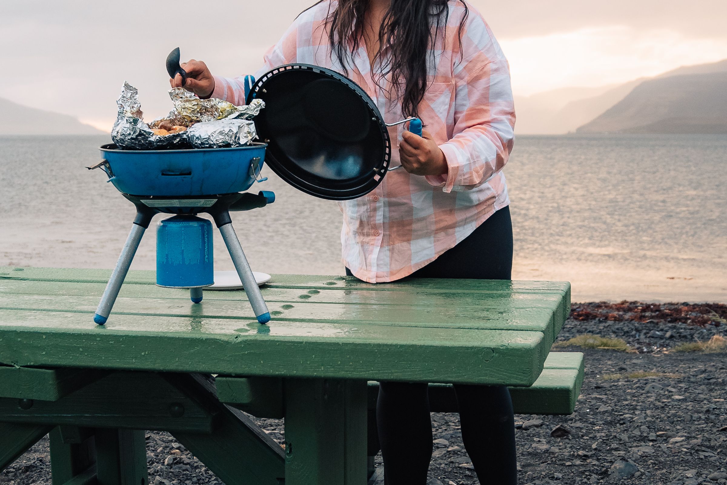 A woman lifts the lid of a portable blue grill cooking foil-wrapped food, on a green picnic table by a lake with mountains.