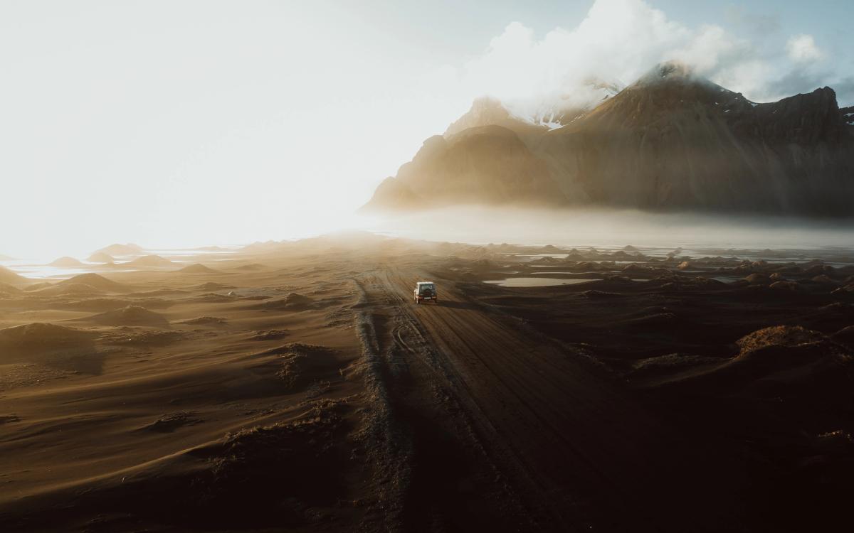 A white van drives on a dirt road through a vast, dark volcanic landscape under hazy sunlight with misty mountains in the distance.