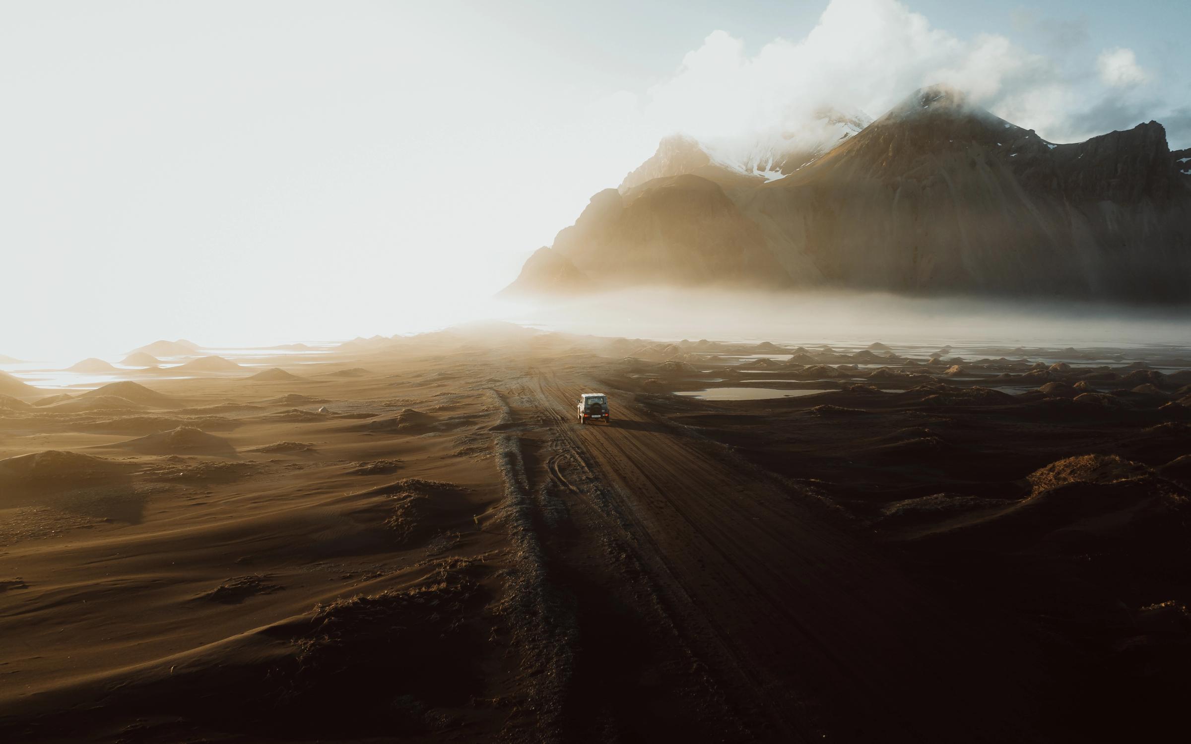 A white van drives on a dirt road through a vast, misty landscape with dramatic mountains under a golden sunrise.