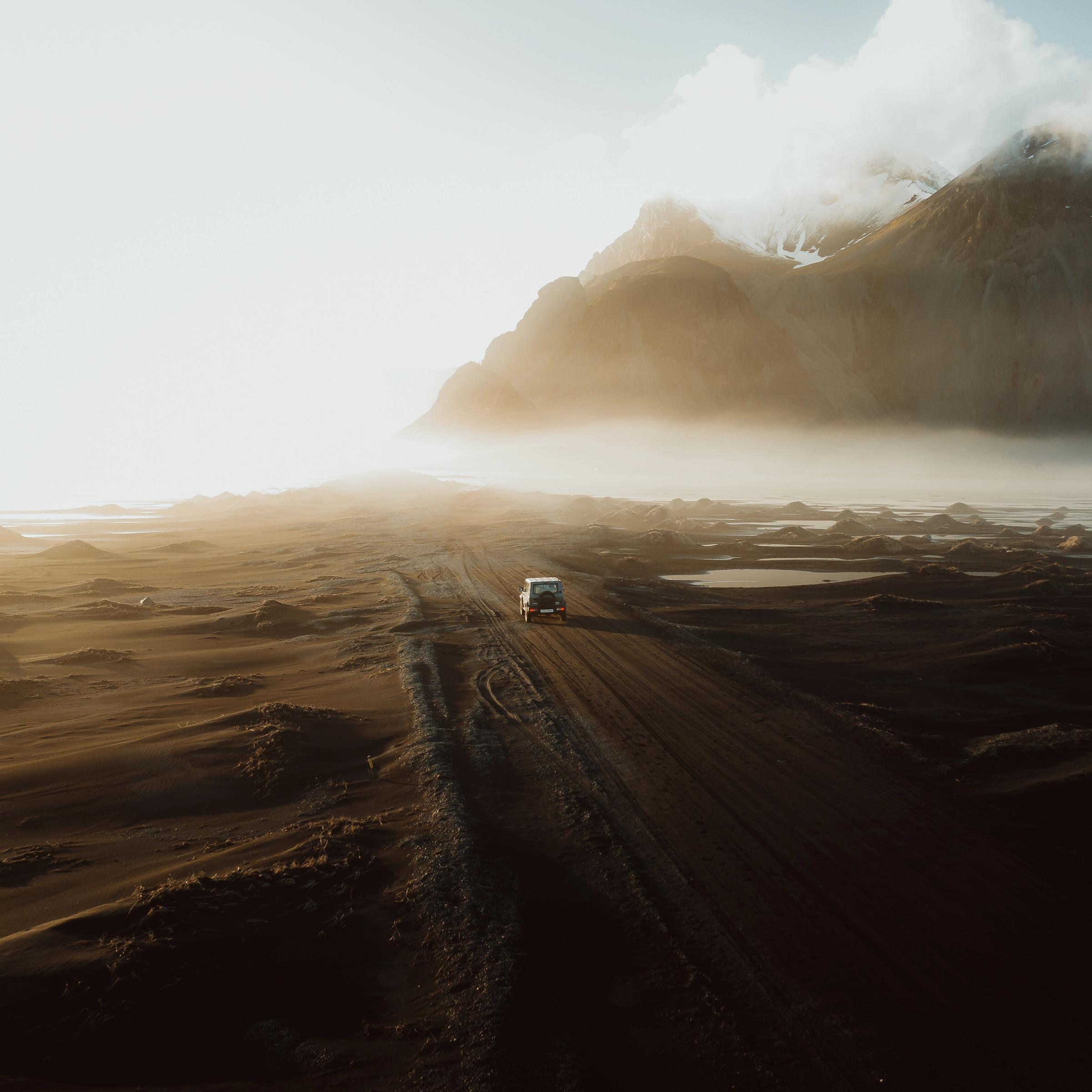 A white van drives on a dirt road through a vast, misty landscape with dramatic mountains under a golden sunrise.