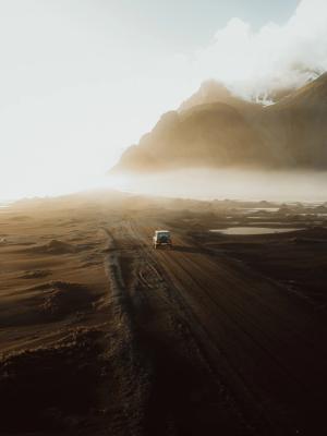 A white van drives on a dirt road through a vast, dark volcanic landscape under hazy sunlight with misty mountains in the distance.