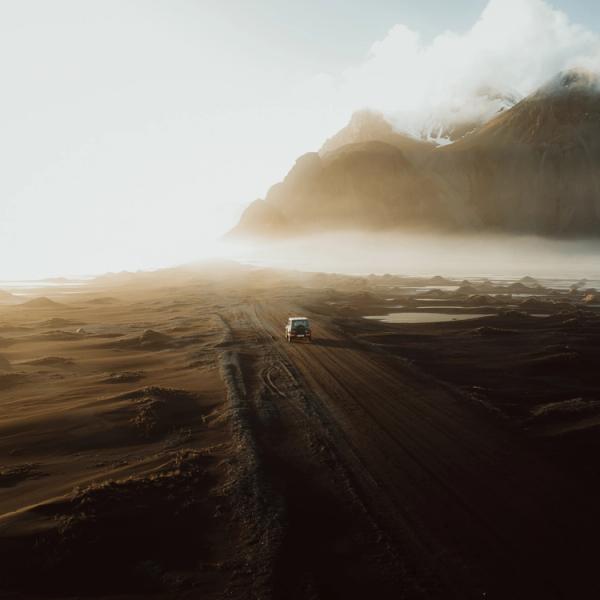 A white van drives on a dirt road through a vast, misty landscape with dramatic mountains under a golden sunrise.