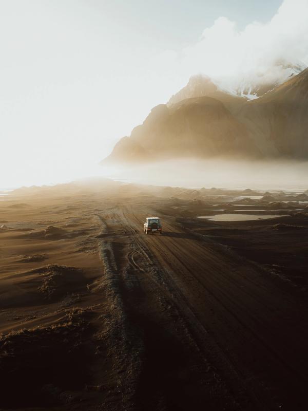 A white van drives on a dirt road through a vast, dark volcanic landscape under hazy sunlight with misty mountains in the distance.