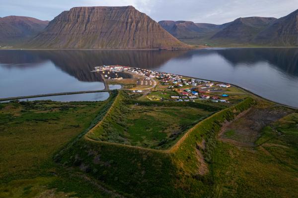 Aerial view of a coastal town on a peninsula, flanked by green grassy earthworks, with a fjord reflecting mountains in the background.