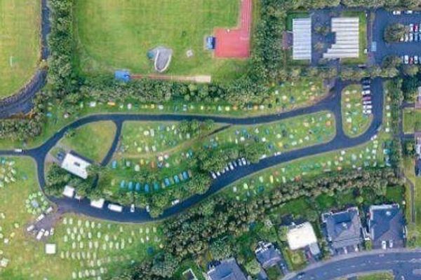 Aerial view of a large campsite with colorful tents spread across green fields, bordered by roads and buildings.