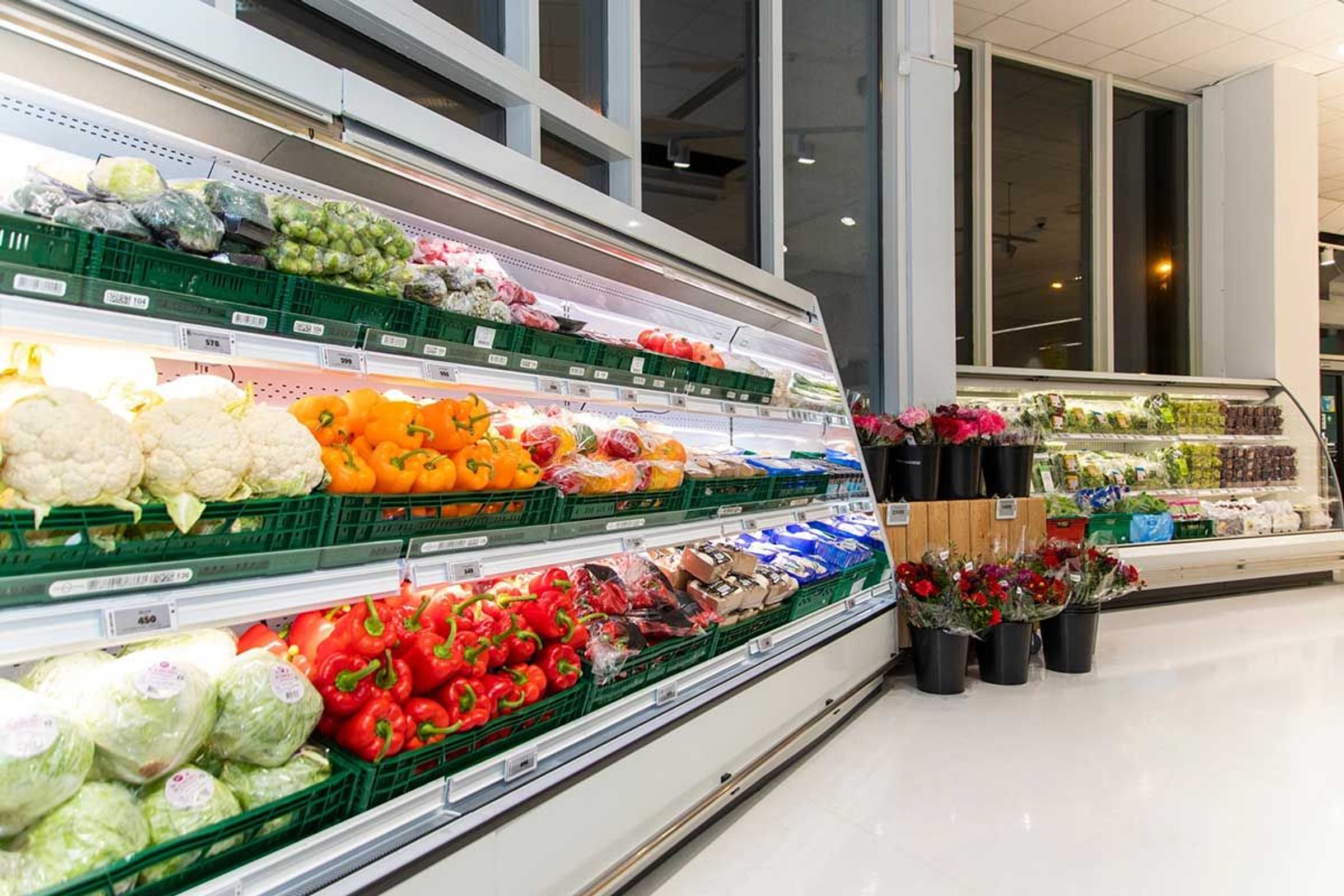 Pasillo de supermercado con vitrinas refrigeradas que exhiben productos frescos como pimientos y coliflor, y ramos de flores.