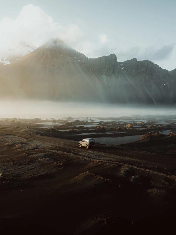 A white SUV drives on a dirt road through a dark, misty landscape with jagged mountains under a golden light.