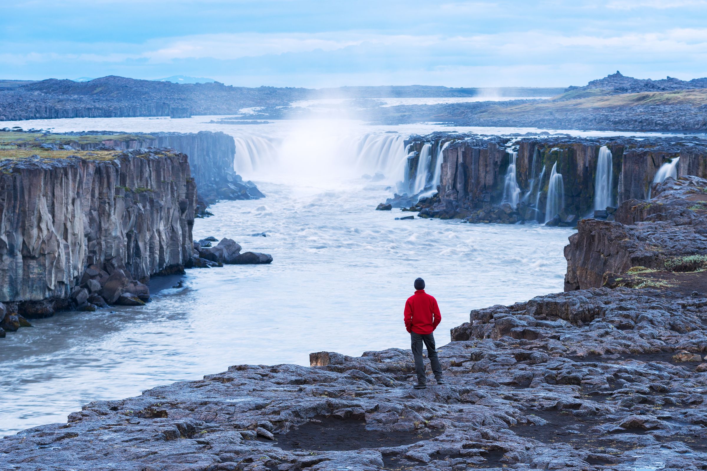 A person in a red jacket stands on a rocky cliff overlooking a wide river with multiple waterfalls.