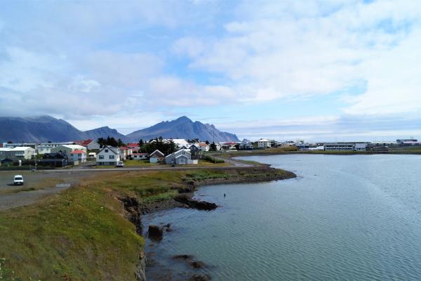 A town with houses along a bay, backed by tall mountains under a cloudy sky.