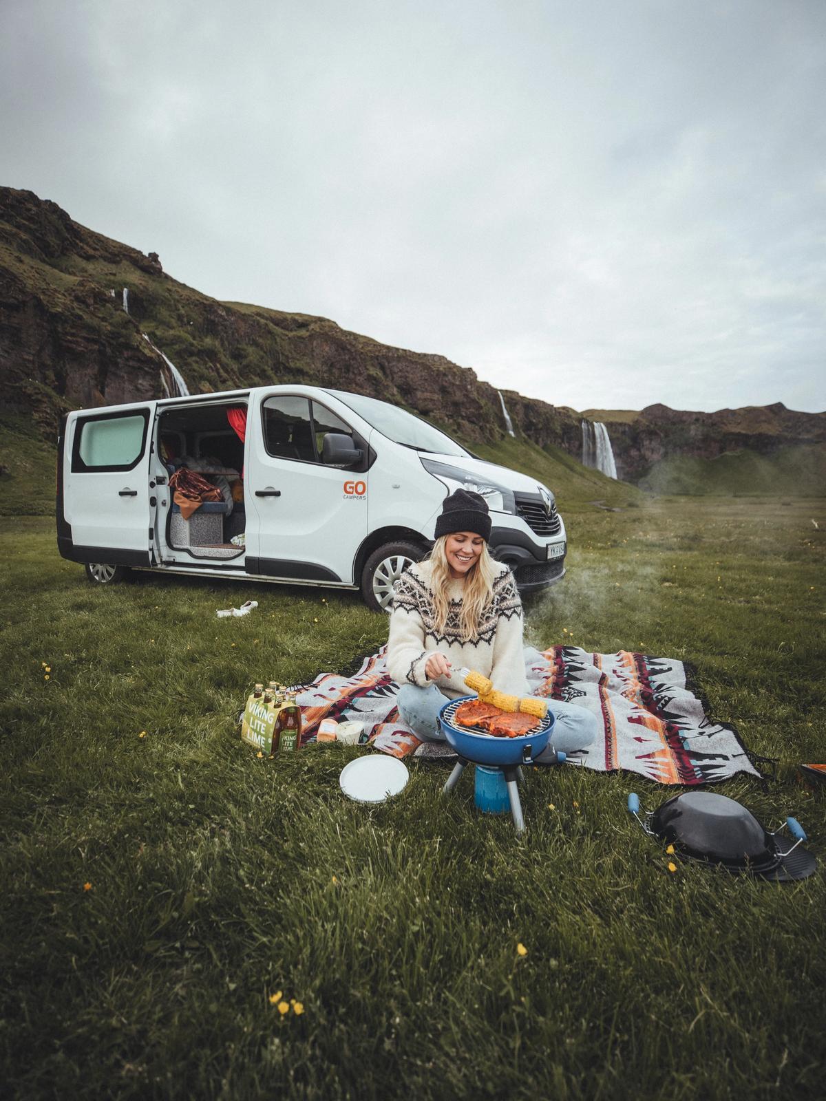 a woman is sitting on a blanket cooking food in front of a van .