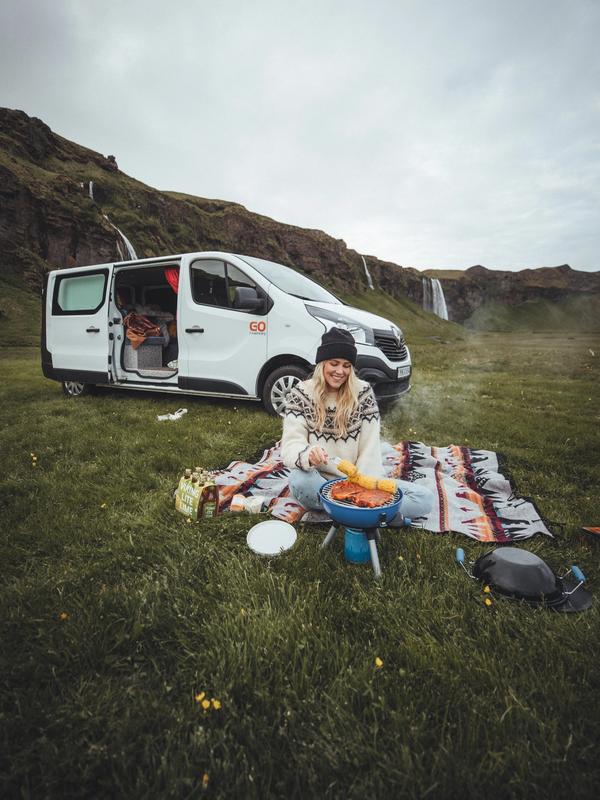 a woman is sitting on a blanket cooking food in front of a van .