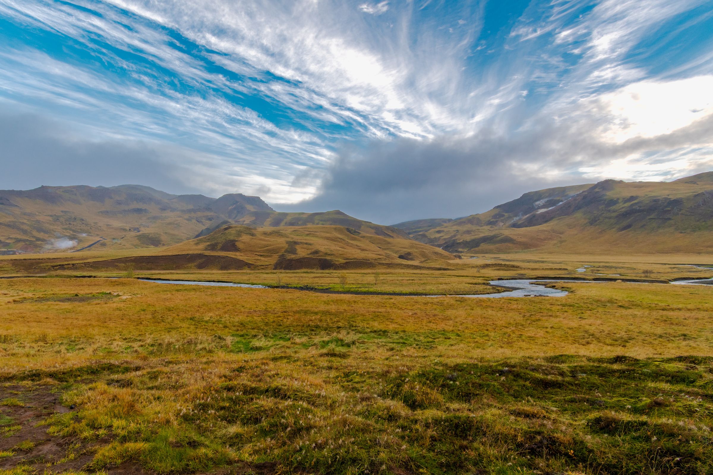 Un valle dorado cubierto de hierba, con un río serpenteante, colinas onduladas y un cielo azul intenso lleno de nubes finas y vaporosas.
