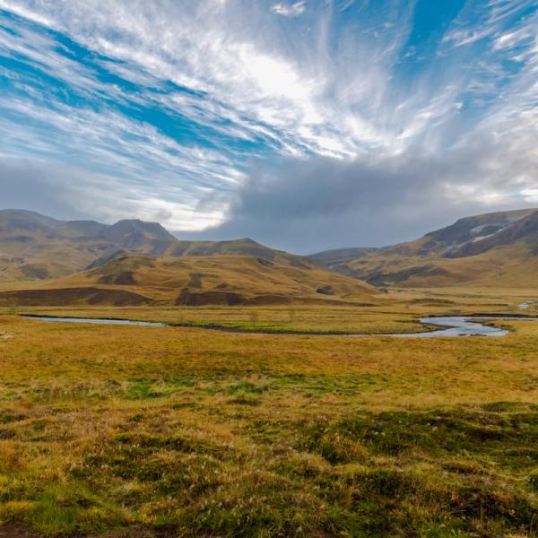 A winding river through a golden grassy valley with rolling hills under a dramatic blue and white sky.