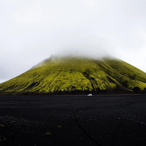 A green mossy mountain with its peak obscured by clouds rises from a dark volcanic plain with a small white vehicle.